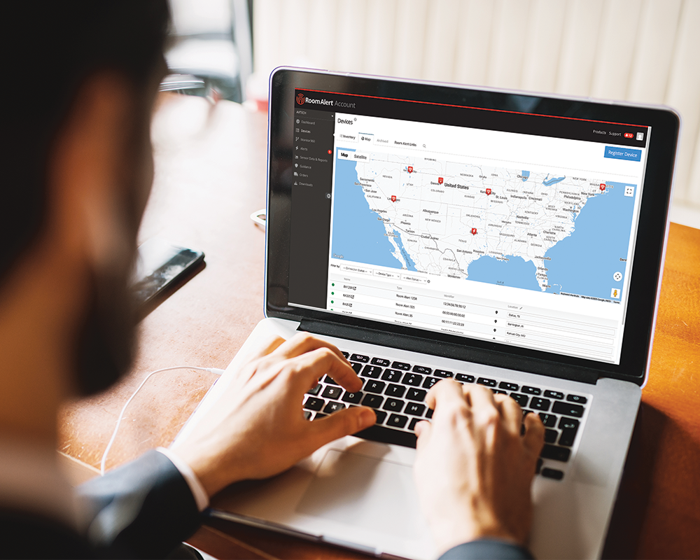 Person using a laptop computer displaying the Room Alert Account monitoring dashboard with a map interface showing multiple monitoring locations across the United States, demonstrating multi-site facility monitoring capabilities.
